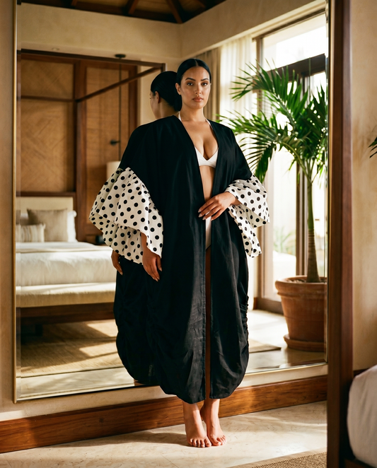 Woman in a black kimono with white and black polka dot sleeves standing in a room with a mirror and plant. by Mesan