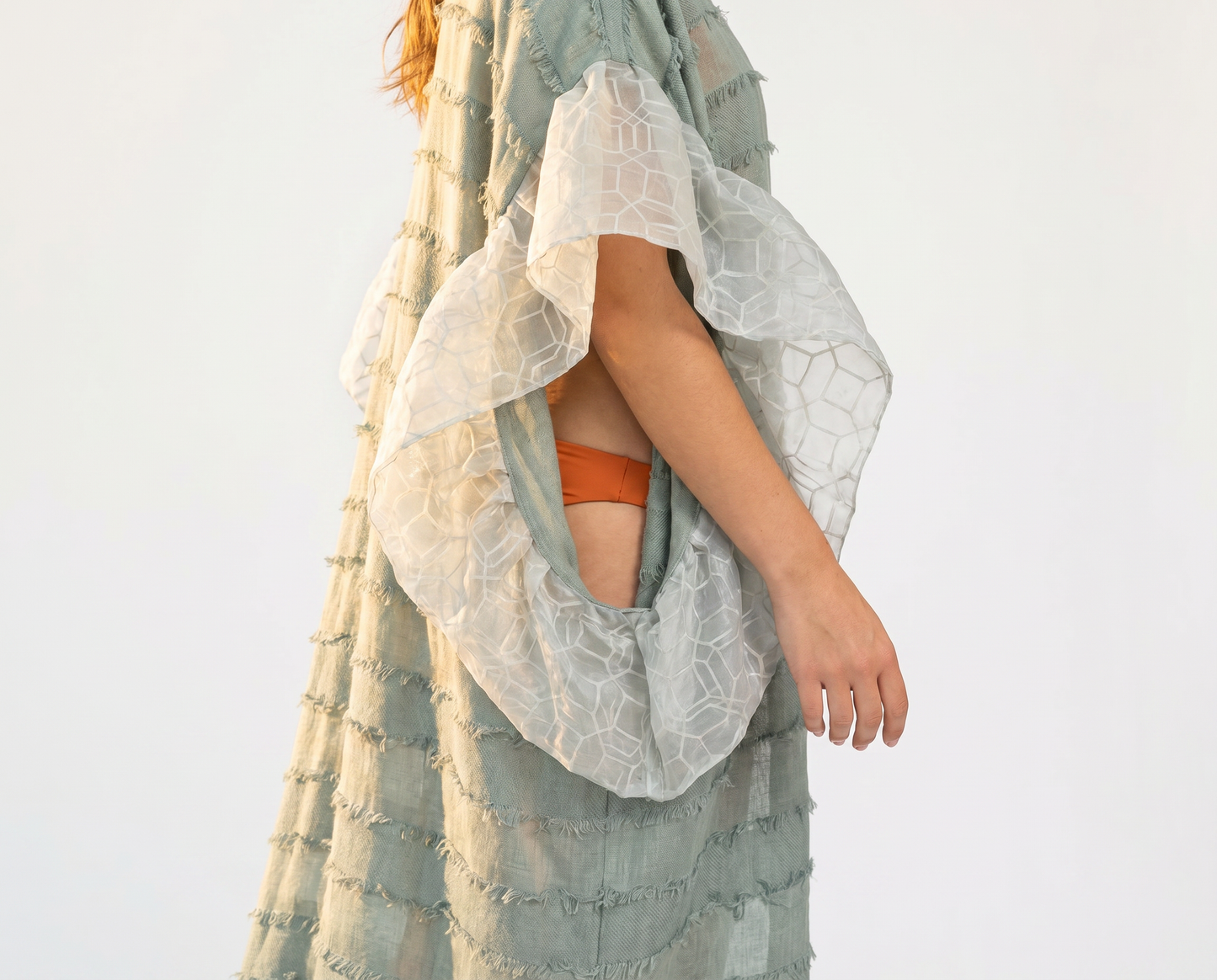 Person wearing Mesan sage green colored kaftan with short striped fringe details and organza ruffles, with an orange bathing suit bottom, white background